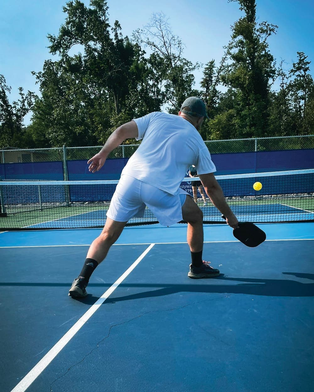 Un joueur de pickleball, vu de dos, frappe la balle en coup droit sur un terrain bleu, en plein soleil