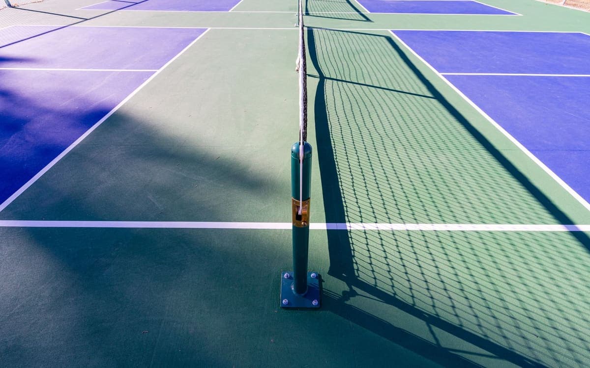 Un filet de pickleball tendu sur son poteau vert, entre des terrains bleus et verts en plein soleil